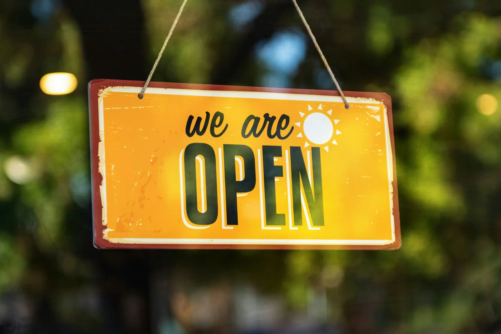 一塊用細繩吊著的黃色招牌，寫著英文 we are open 和一個小太陽圖案，背景是模糊的綠色樹葉與光點